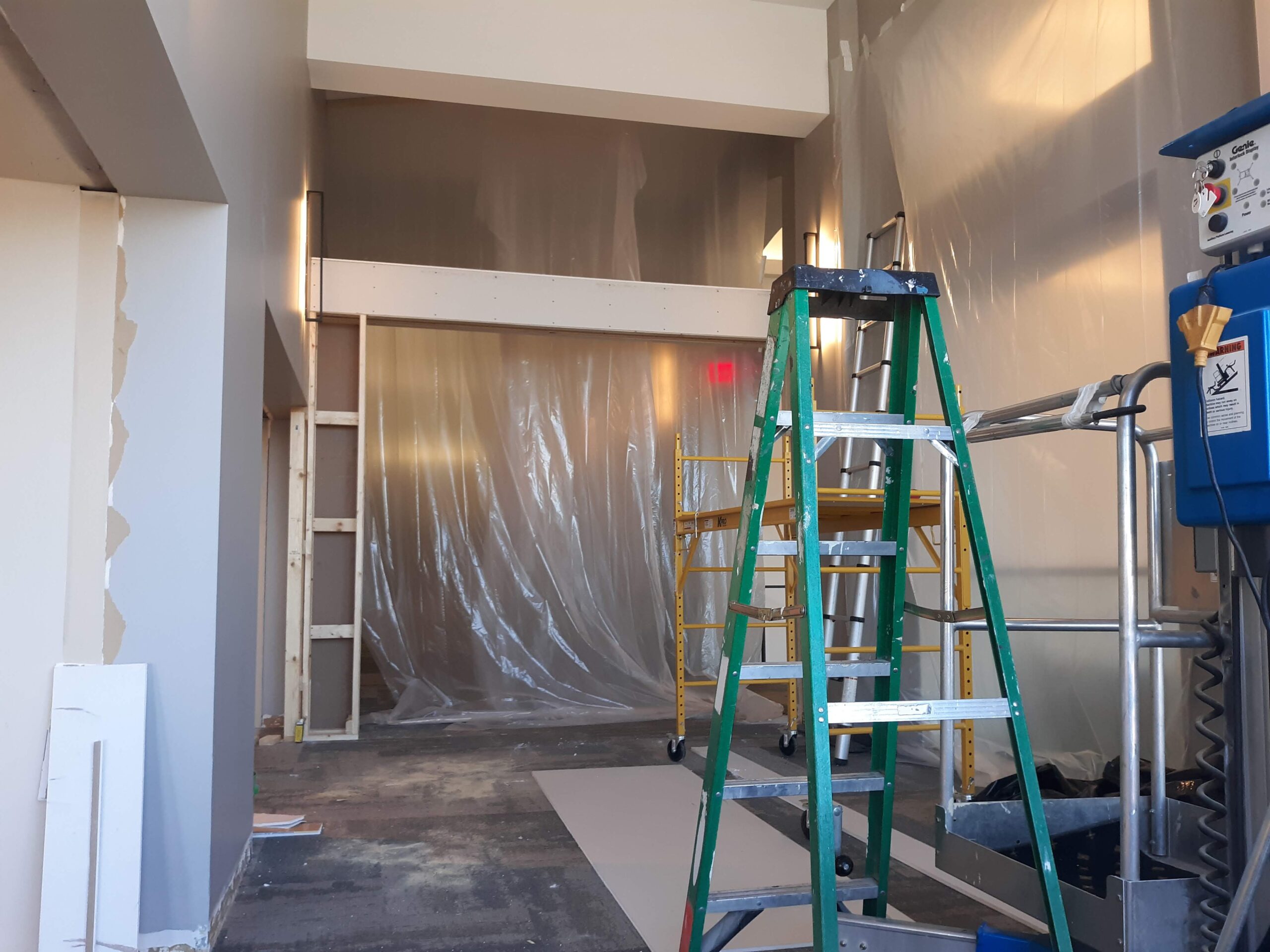 A commercial space under construction with a stepladder and scaffolding in the foreground, and a large doorway covered in plastic sheeting in the background.